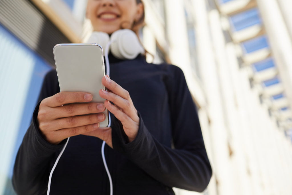 Checking sms on gadget during jogging Close-up of smiling sporty jogger in black jacket wearing headphones on neck checking sms on gadget during jogging in city