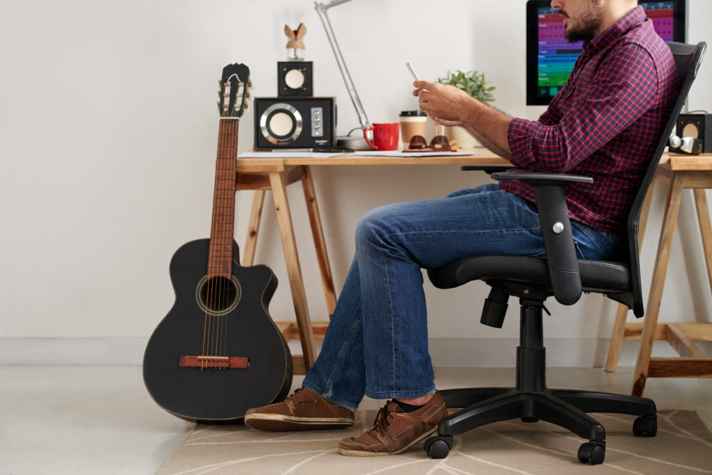 Man using his smartphone Young casual man sitting on chair near the table with computer and guitar and using his smartphone while working at sound studio