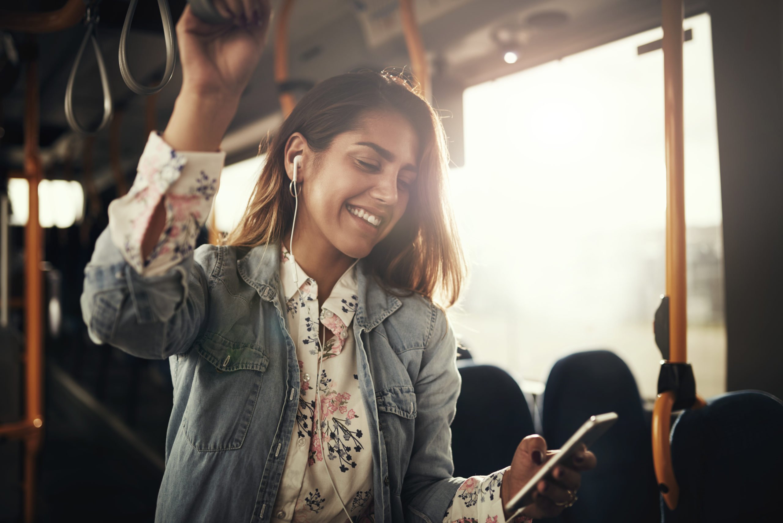 Young woman smiling while standing by herself on a bus listening to music on a smartphone